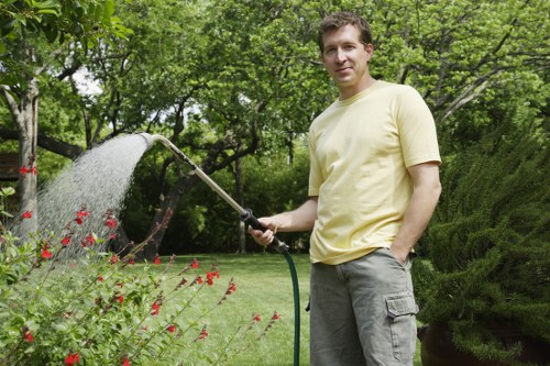 A man standing in a well-maintained garden during daytime, holding a garden hose and watering flowering plants with red blossom clusters. The garden features a neatly trimmed grassy lawn, bordered by a variety of shrubs and trees with lush green foliage, providing a natural and inviting outdoor environment. In the background, tall trees cast dappled shade over the garden, while a dense hedge creates a sense of privacy. The scene captures a peaceful and tidy outdoor space typical of residential gardening in Greenwich, with natural sunlight illuminating the scene and emphasizing the healthy, vibrant plant life. This image illustrates outdoor maintenance activities that a professional gardening service like Garden Maintenance Greenwich may undertake to keep gardens attractive and thriving.
