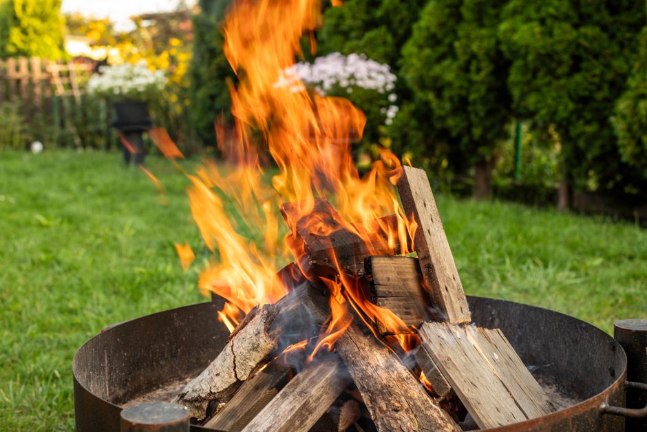A large bonfire burning outdoors during night time, with bright orange flames consuming a heap of dry twigs, branches, and leaves. The flames produce intense heat and light, illuminating the surroundi